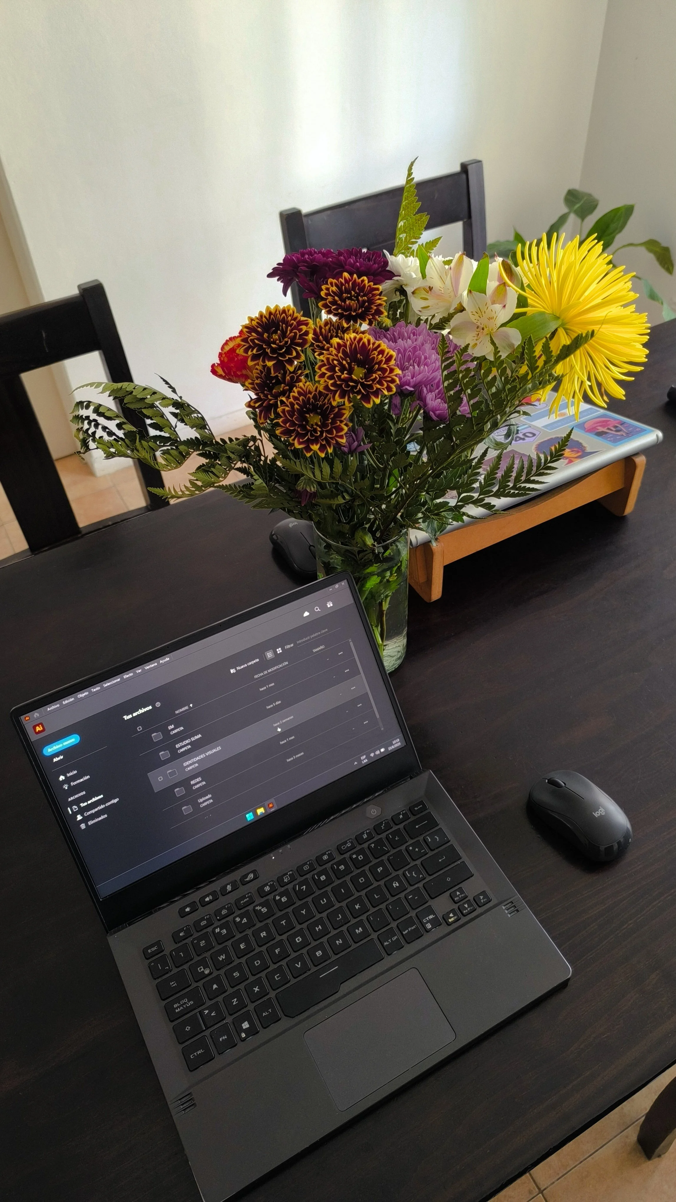 Mesa de oficina con una laptop abierta, un mouse y un florero con flores de varios colores, sobre una bandeja de madera.