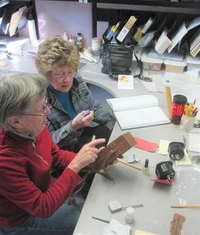 Two women sitting at a table in an office or workspace, examining a booklet, with various office supplies and shelves in the background.
