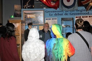 Group of people gathered around a display case at an exhibit, some wearing colorful jackets and hoodies, with framed pictures and text in the background.