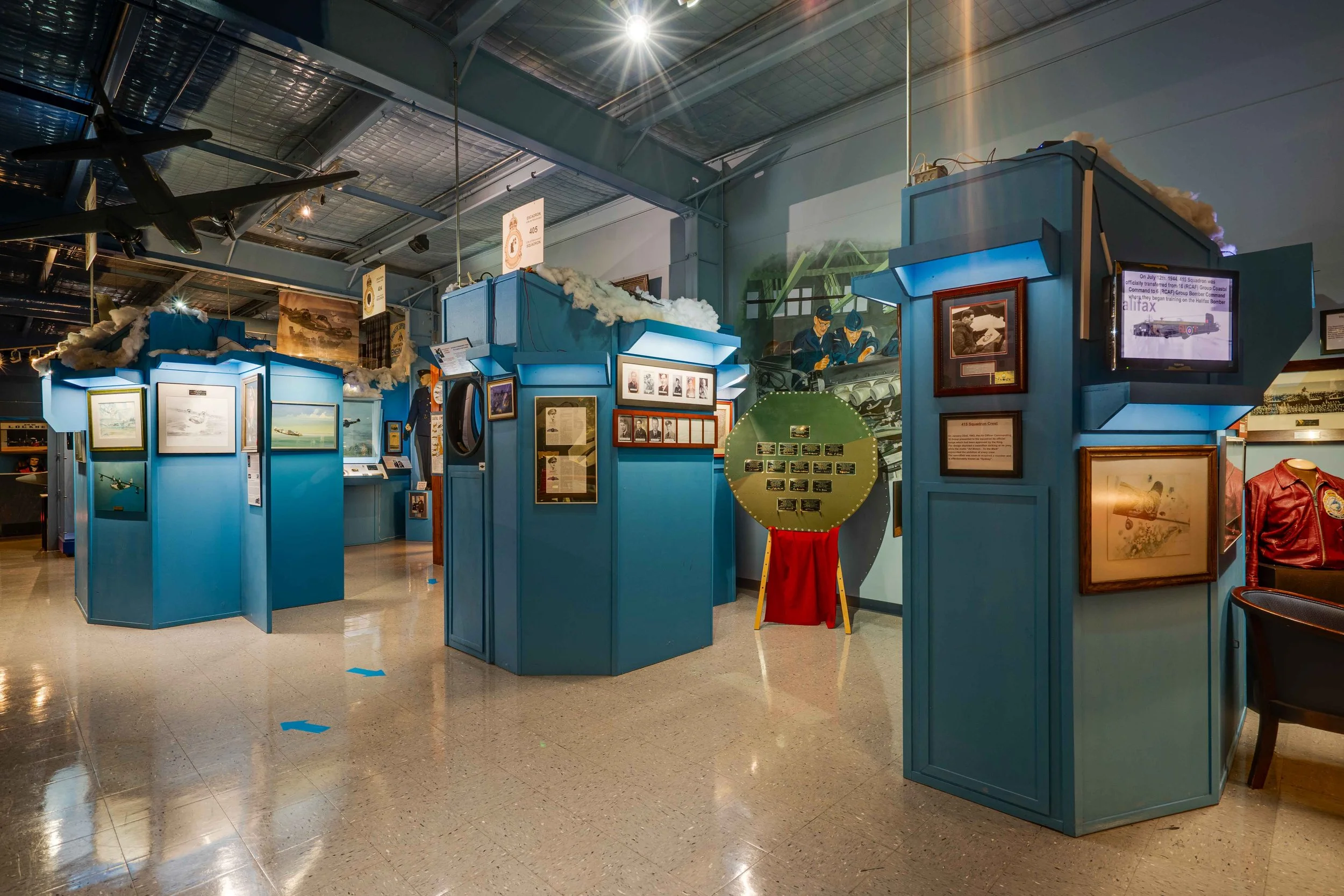 A museum exhibit with blue display booths showcasing aviation history, model airplanes, photographs, and memorabilia, with a painted mural and airplane models hanging from the ceiling.