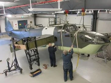 Two workers inspecting or maintaining a military aircraft inside a hangar, with equipment around them.