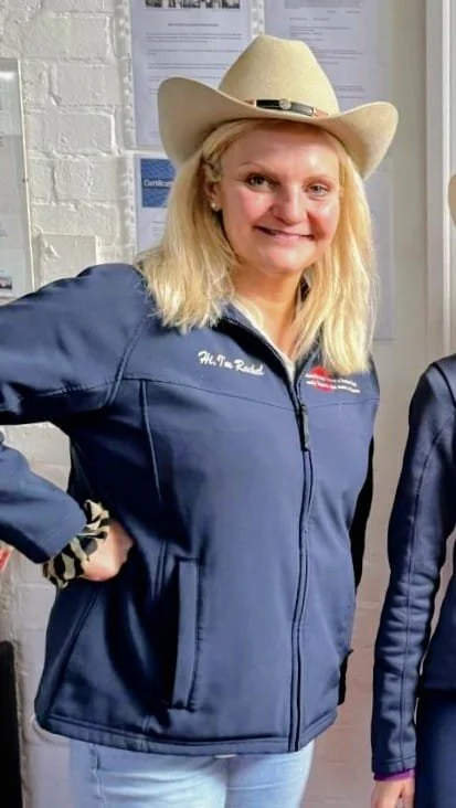 A woman wearing a beige cowboy hat and a navy blue jacket with initials smiling and standing indoors.