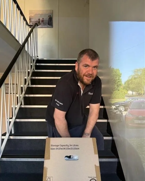 A man with a beard sitting on stairs inside a building, holding a cardboard box with storage information, near a large window showing parked cars and trees outside.