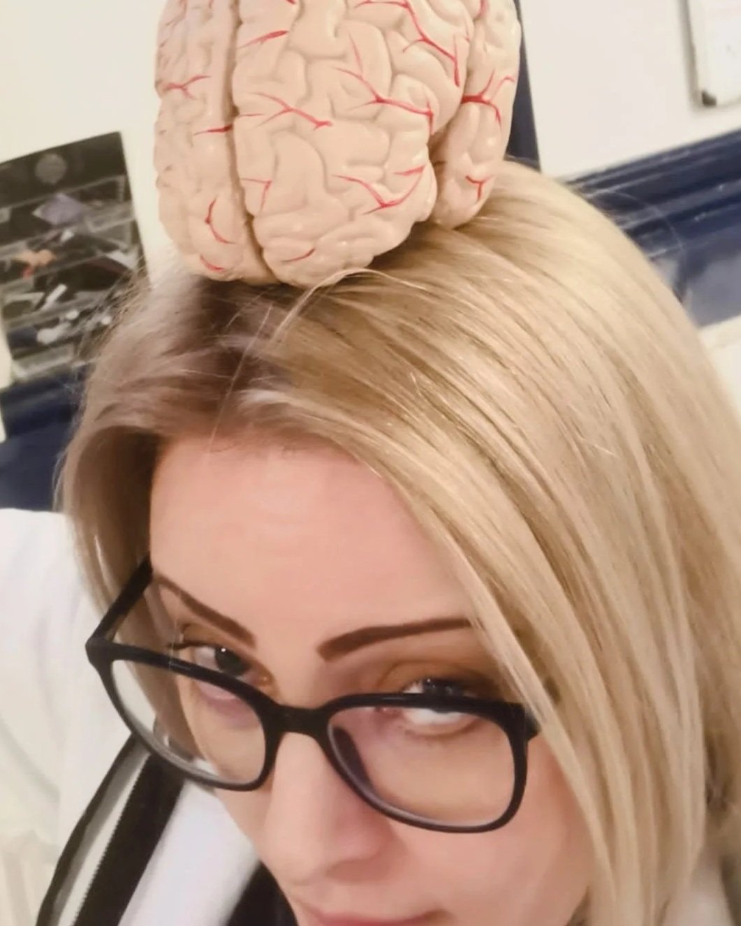 A woman with blonde hair and glasses is seen from above, with a model of a human brain placed on her head.