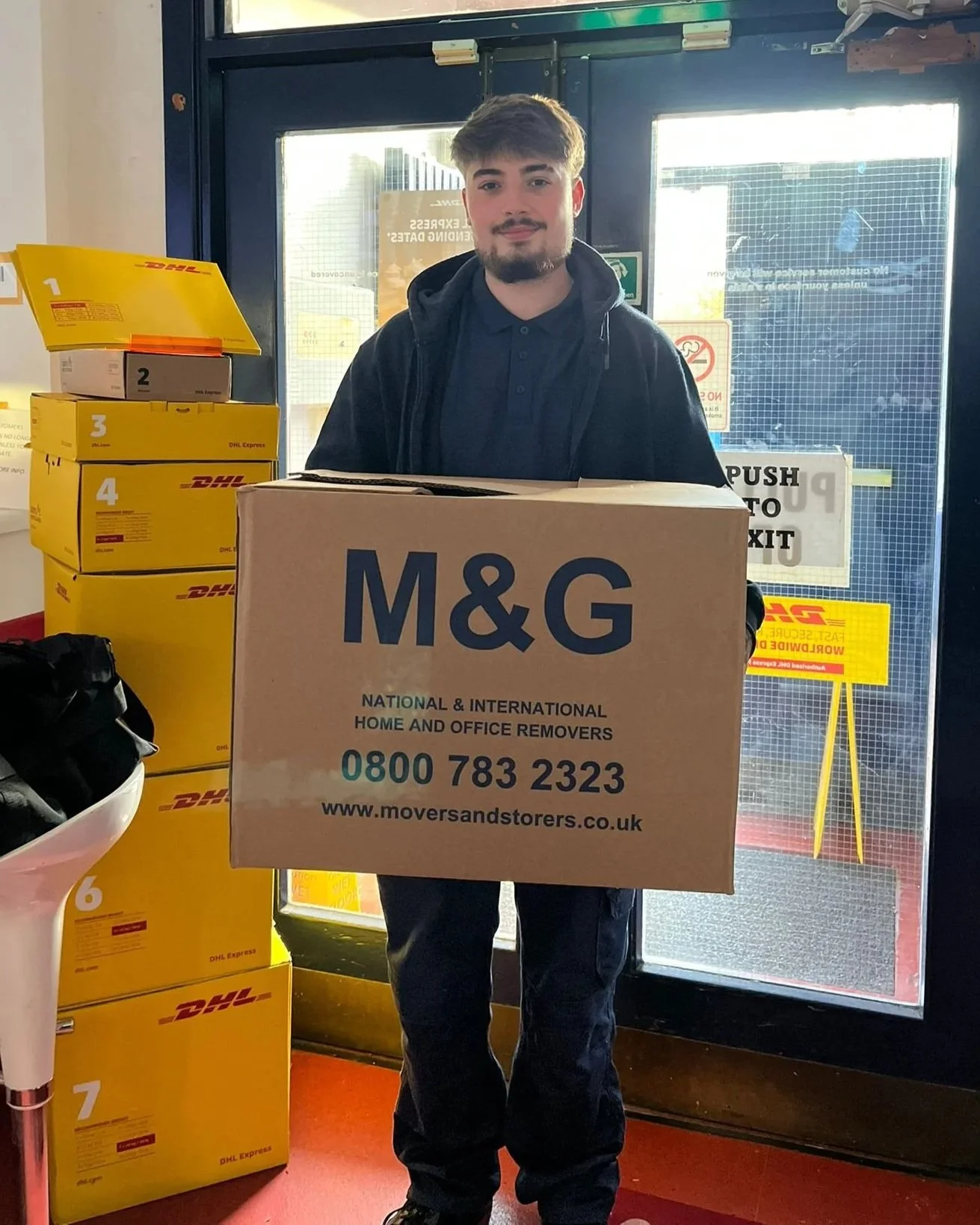 A young man with dark hair and a beard holding a large M&G moving box inside a DHL store, with several DHL boxes stacked around him and a door with signs in the background.
