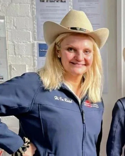 Woman wearing a beige cowboy hat and a blue jacket, smiling indoors.