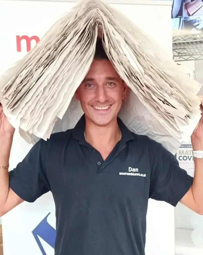 A man smiling and holding a large stack of papers or newspapers above his head, creating a tent-like shape.