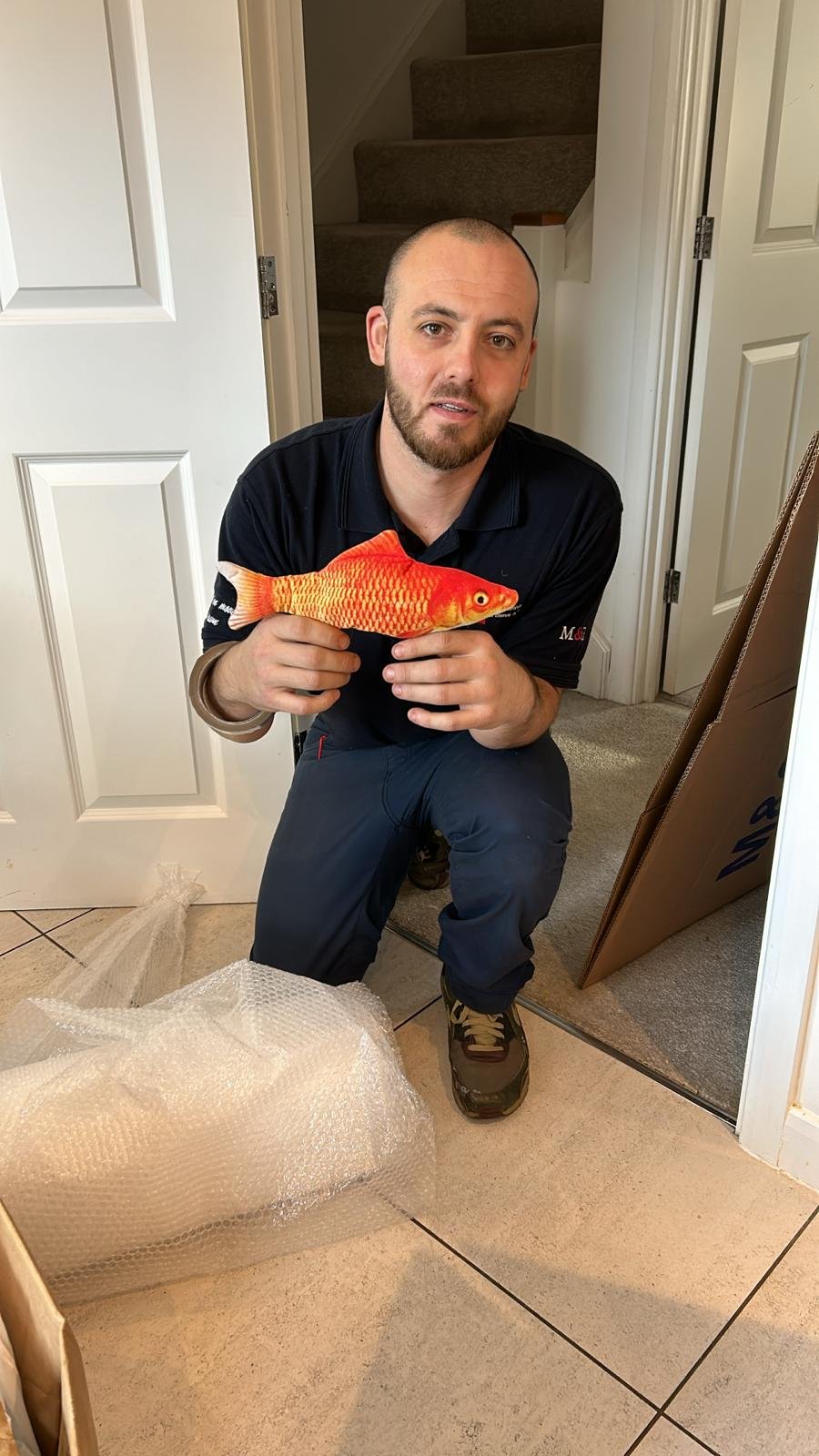 A man crouching indoors, holding an artificial orange and yellow fish, next to a pile of bubble wrap on the tiled floor, with a doorway and carpeted stairs in the background.