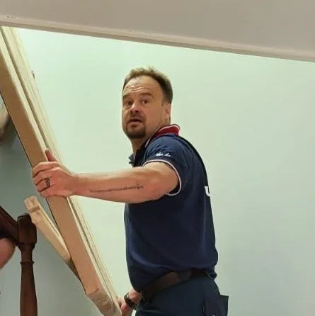 A man with short hair and a beard looking surprised or confused, holding a large wooden plank while standing indoors.