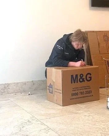 A woman with blonde hair in a ponytail, wearing a black jacket, is bending over and writing on a large cardboard box labeled 'M&G' and 'Overseas & Logistics' in a room with tiled flooring and a white wall, with a small piece of paper or trash on the floor.
