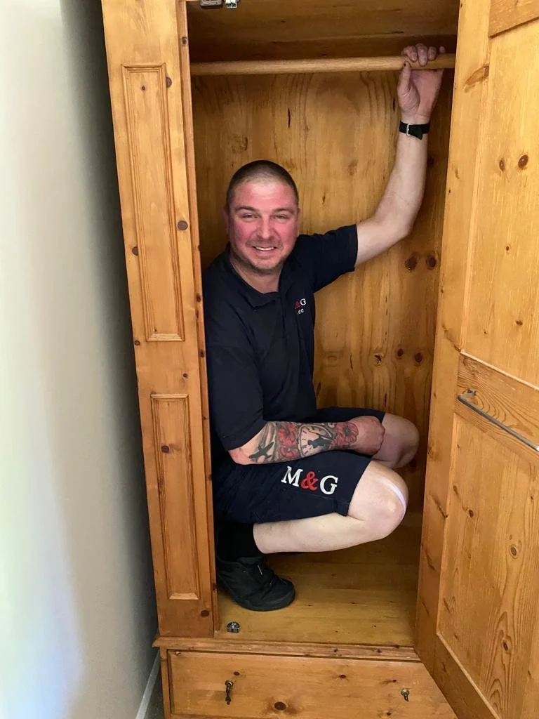 A man is squatting inside a small wooden closet, smiling, with one arm raised and gripping the top of the closet. He has short hair, a tattoo on his arm, and is wearing a black shirt and shorts with a logo.