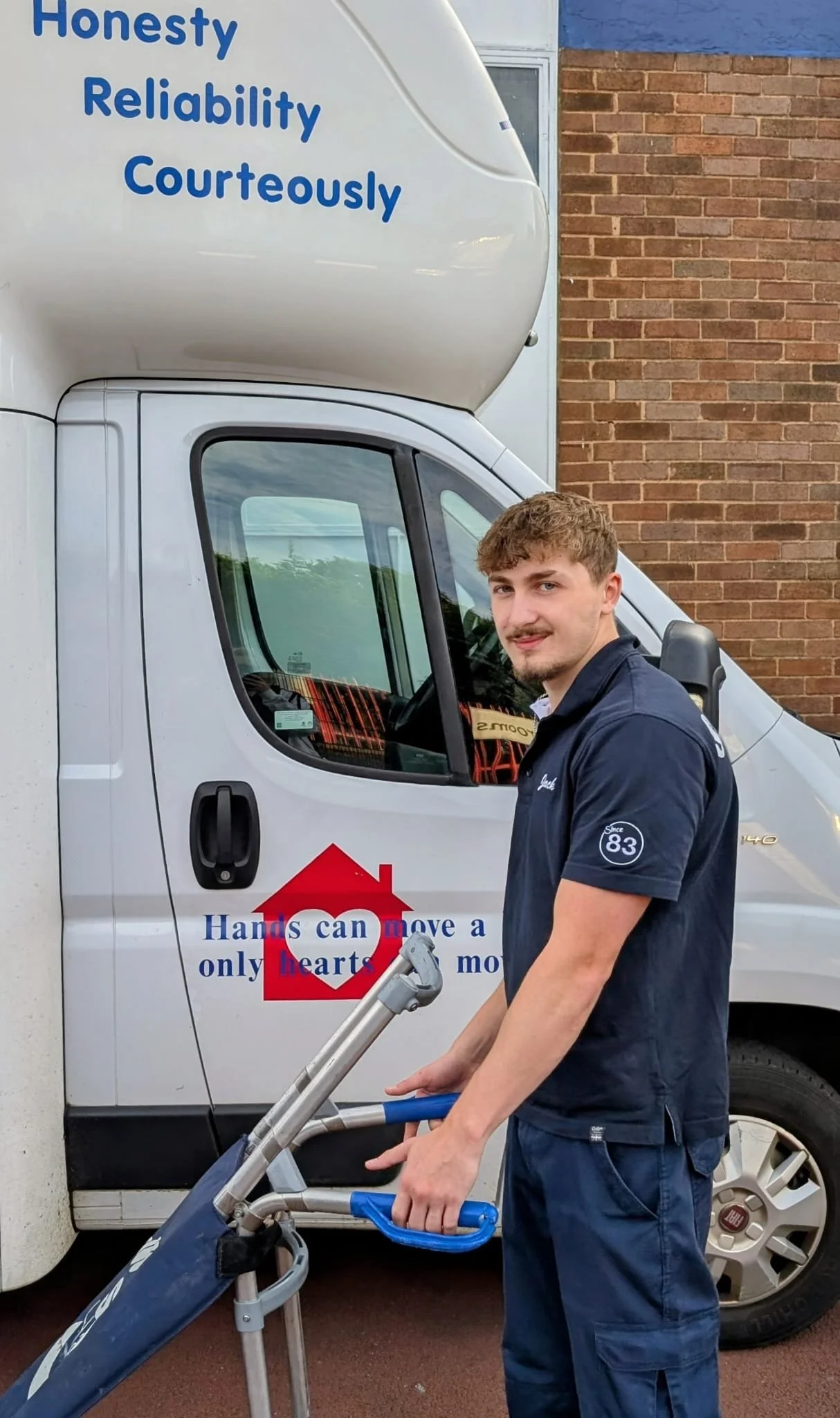 A young man in a navy uniform standing next to a moving company van holding a hand truck. The van has an inspirational quote about honesty, reliability, and courtesy, and a red heart logo with the words 'Hands can move a heart only'. The background features a brick wall.