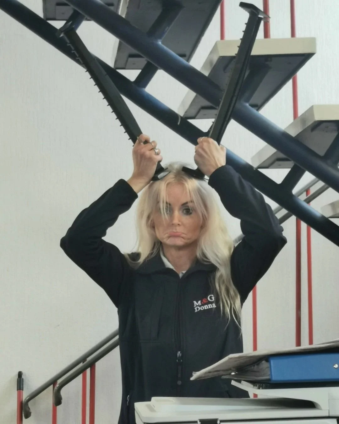 A woman with long blonde hair, wearing a black jacket with a logo that says 'Donna', holds a staircase rail above her head with a serious expression.