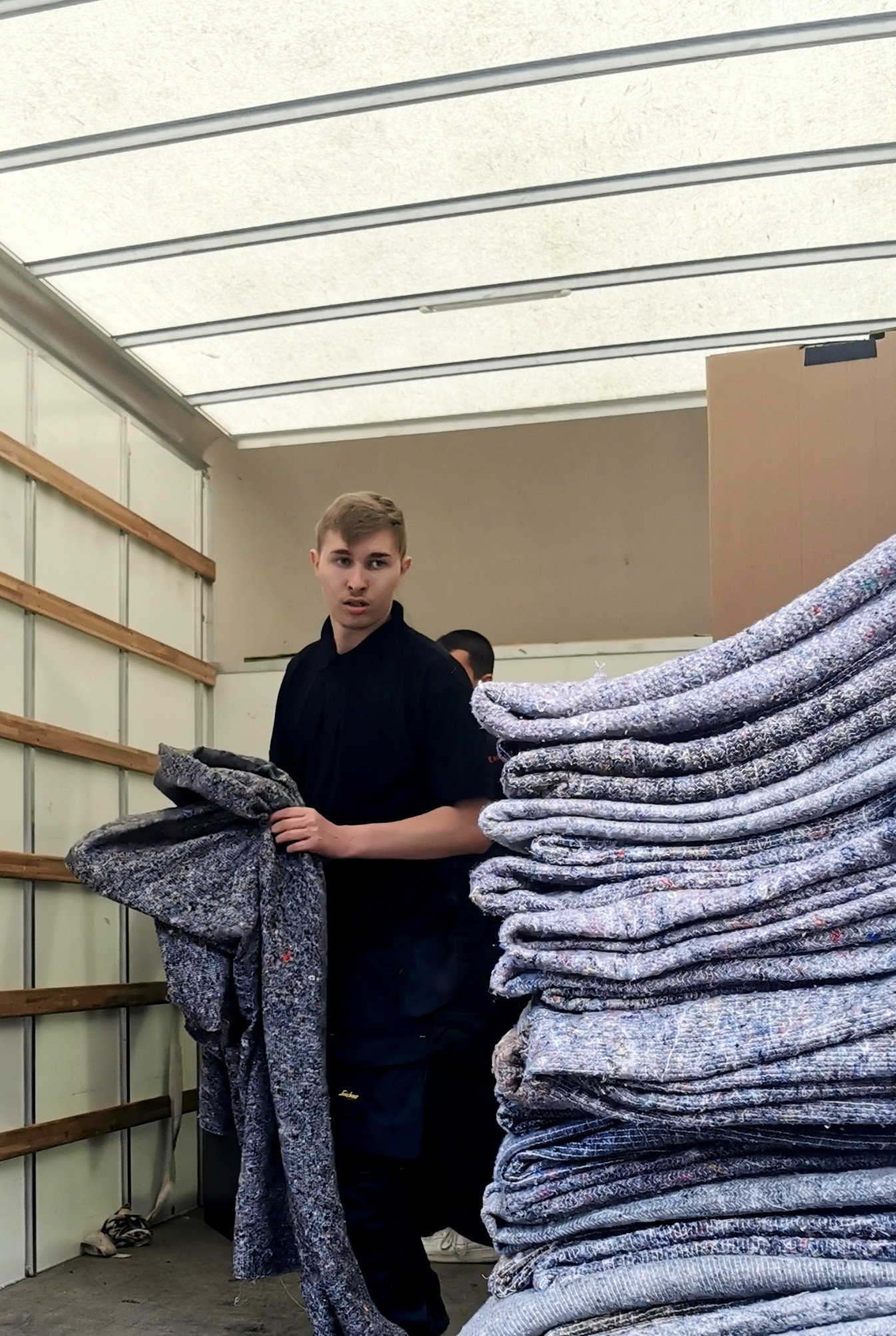 Young man in a black shirt standing in a warehouse with stacks of folded patterned blankets or fabric.