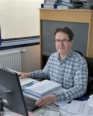 A man wearing glasses and a checkered shirt sitting at a desk with a computer, stacks of paper, and a book in an office.