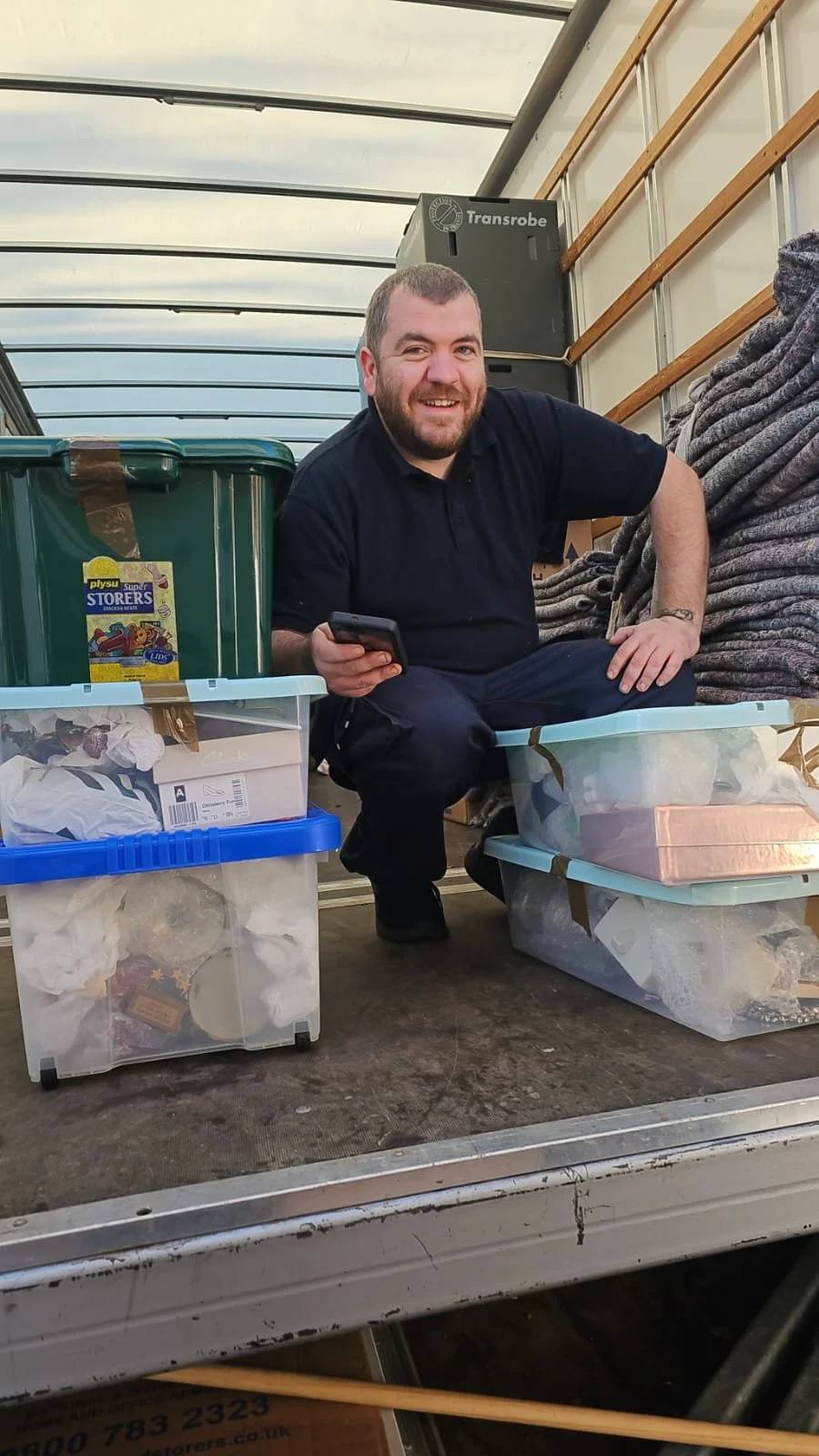 A man with a beard and short hair smiling while squatting in the back of a moving truck, holding a smartphone. The truck contains plastic storage bins filled with various items, a large green storage container, and a pile of gray ropes or fabric on the side.