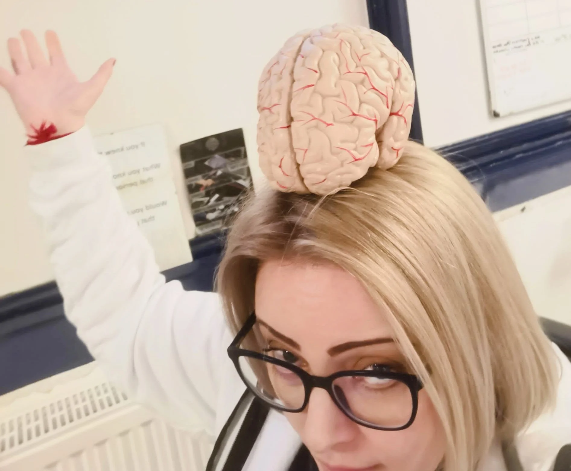 A woman with glasses and blonde hair posing with a model of a human brain on her head in an office.
