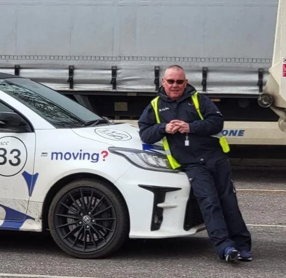A man in a black jacket and safety vest leaning against a white vehicle that has text reading 'moving?' on the side, parked on a road with a large truck in the background.