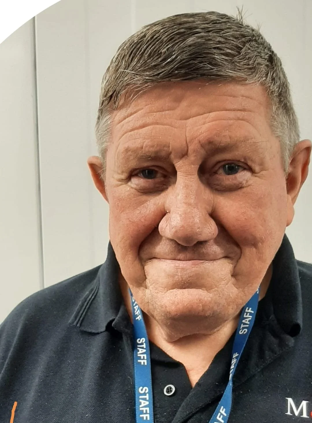 Close-up of a smiling middle-aged man with short, gray and brown hair, wearing a black polo shirt and a blue lanyard with the word 'STAFF' printed on it.