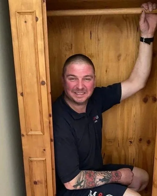 A man with tattoos on his left arm sitting inside a wooden closet, smiling, with his right arm resting on the top of the closet door frame.