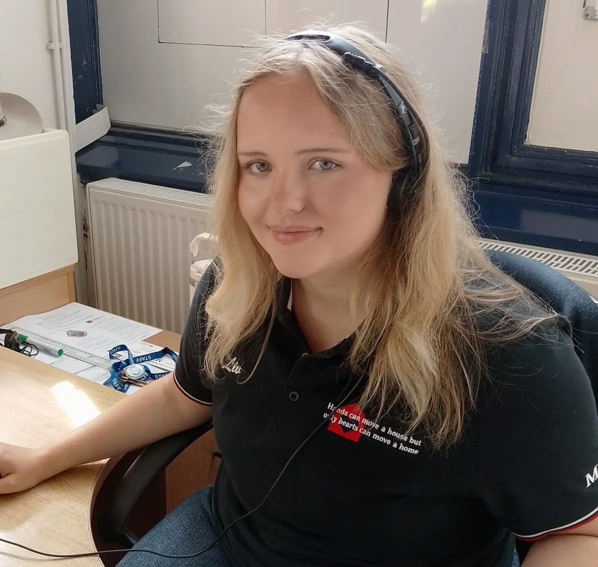 A woman with light blonde hair wearing a black polo shirt with a Red Cross logo and a headset, sitting at a desk in an office.