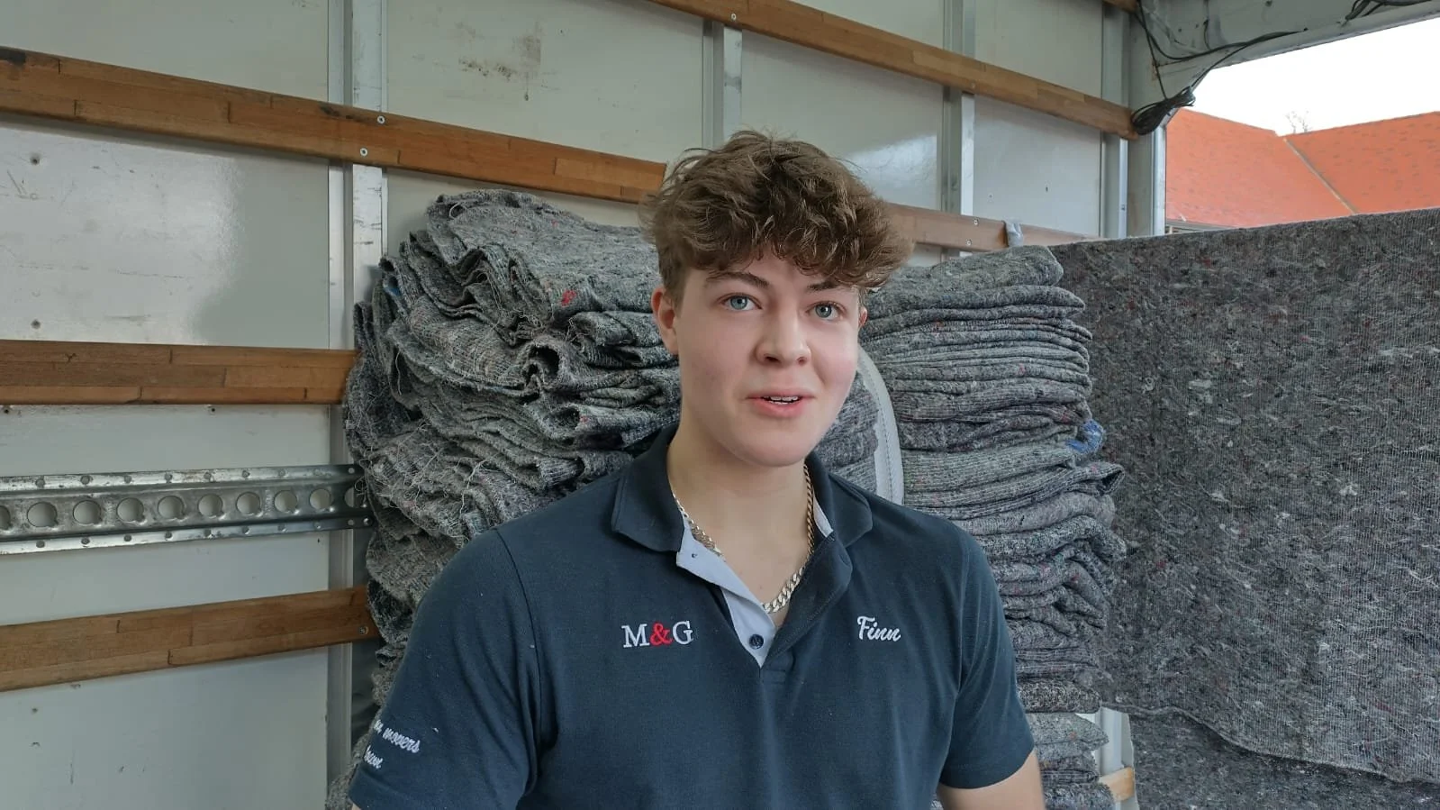 A young man with curly brown hair and blue eyes, wearing a dark blue polo shirt with embroidered text, standing in front of a large stack of grey fabric or carpeting inside a storage unit or warehouse.