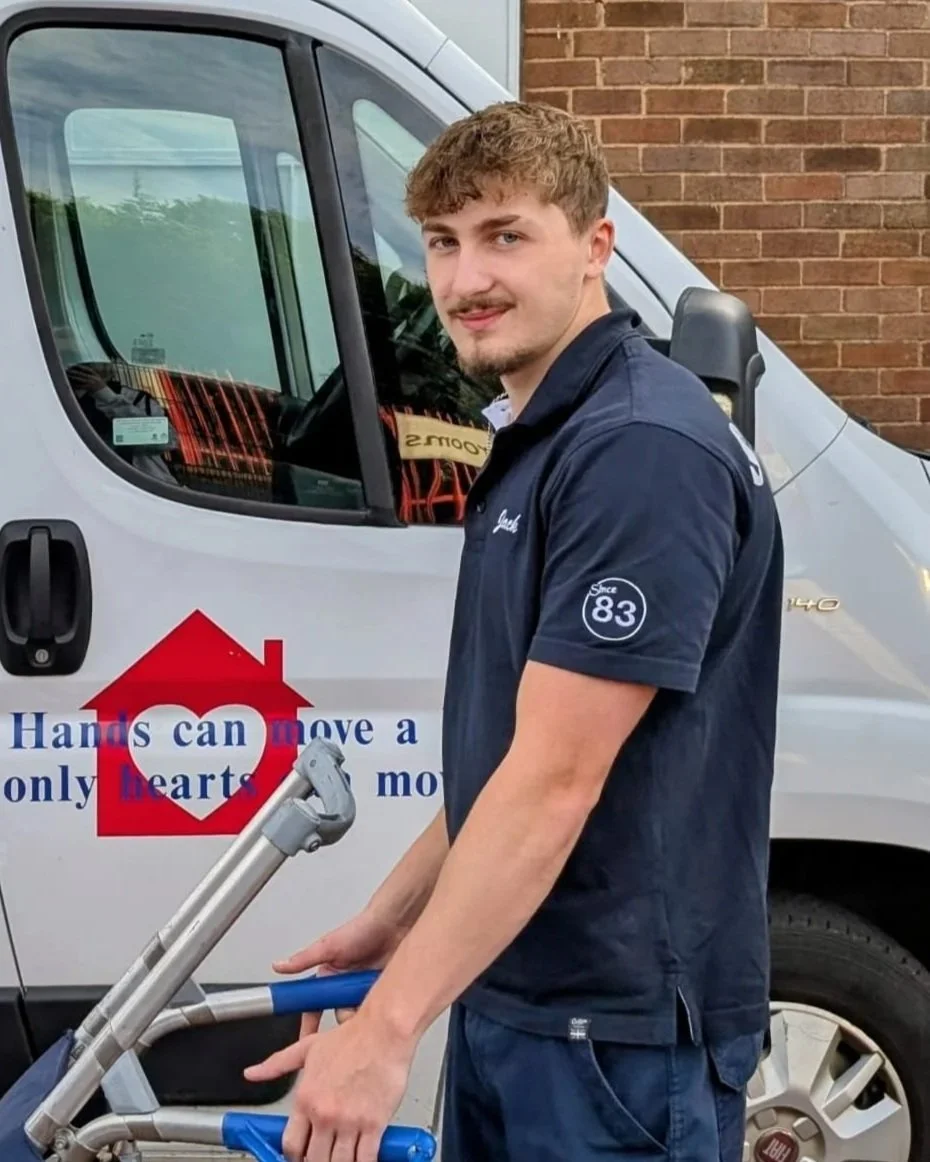 Young man with light brown hair and a beard standing beside a white delivery van, holding a hand truck, with a brick wall in the background.