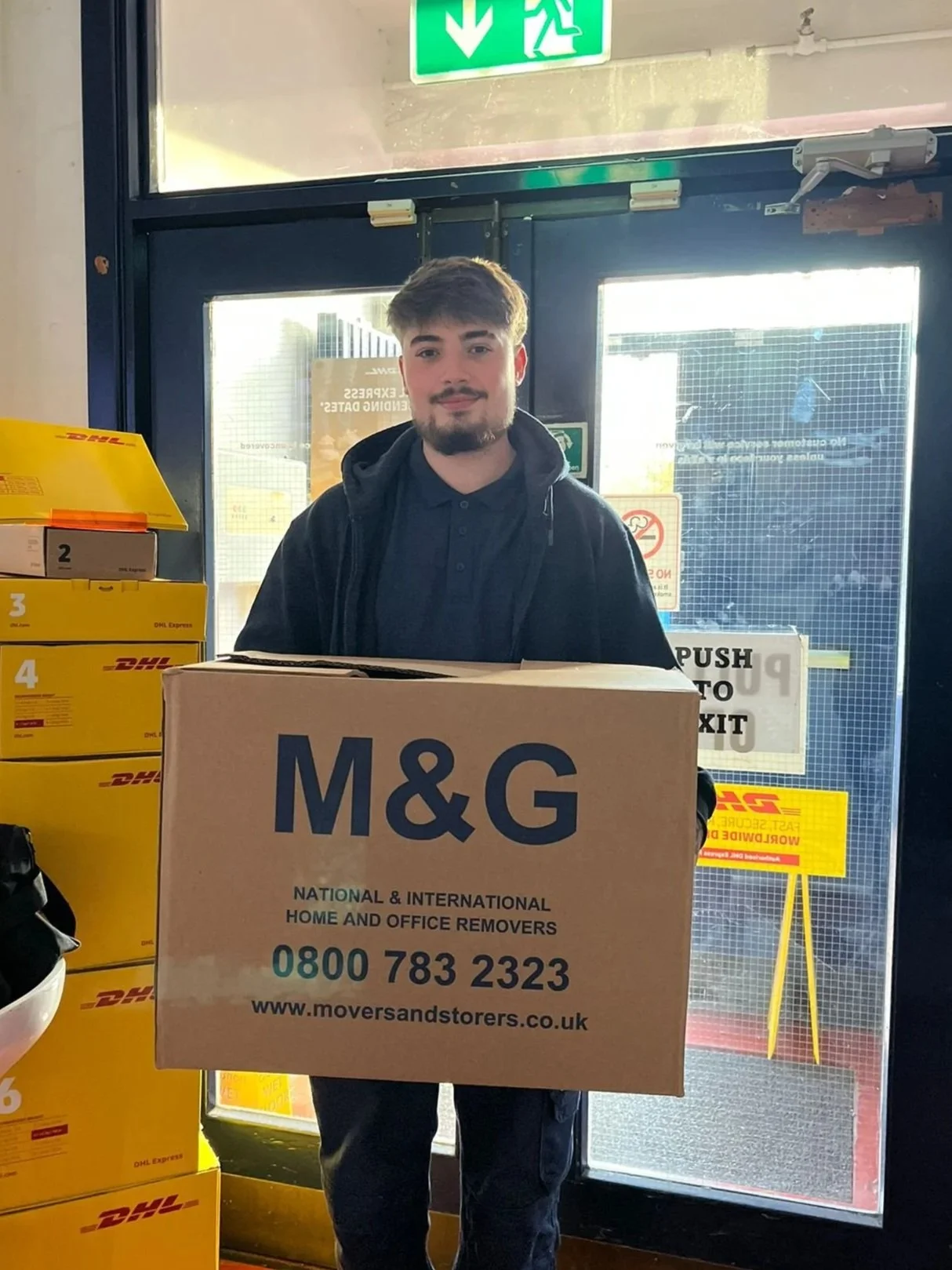 A young man with a beard and mustache holding a large cardboard box with 'M&G' and contact information outside a store with a glass door. There are yellow DHL packages stacked nearby and an exit sign above.