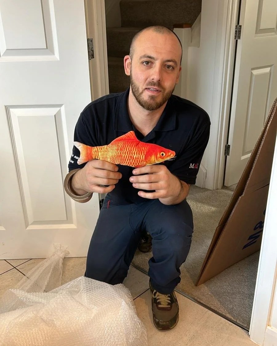 A man kneeling indoors, holding a large, realistic-looking orange and yellow fish plush toy, next to cardboard boxes and bubble wrap.