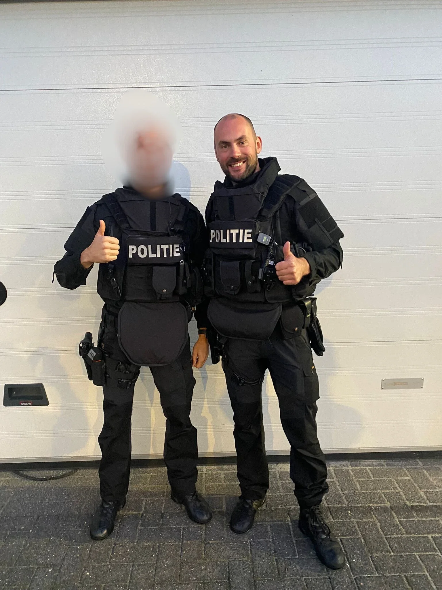 Twee Nederlandse politieagenten in uniform poseert voor een witte garagedeur, allebei geven ze een duim omhoog.