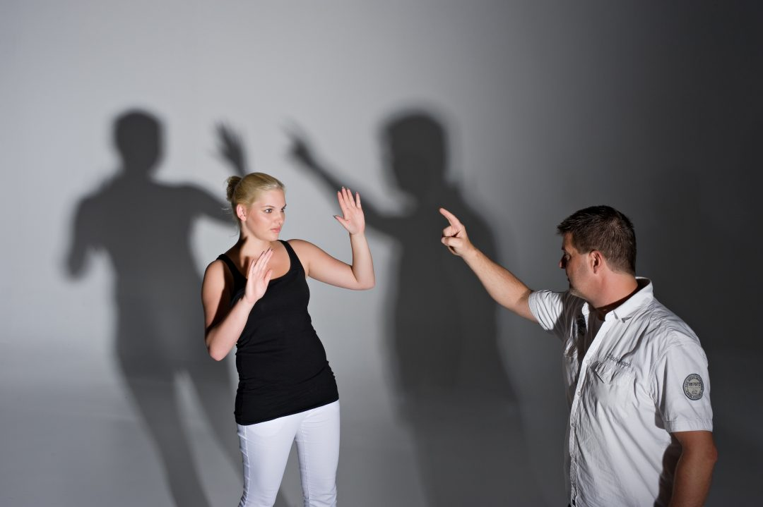 Man wijst naar jonge vrouw die haar handen omhoog houdt, terwijl hun schaduwen op de muur achter hen elkaars bewegingen weerspiegelen.