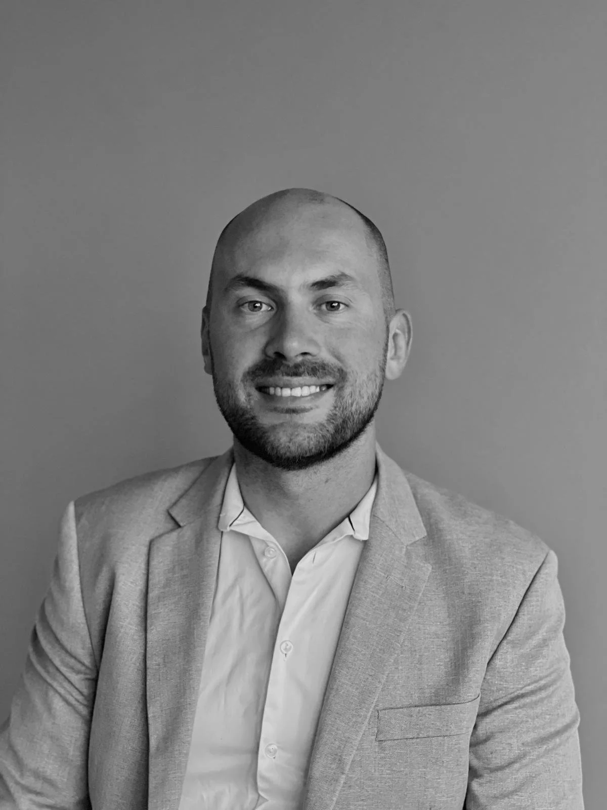 Zwart-wit portret van een man met een baard, korte haar en een lichte blouse, gekleed in een lichtgrijze jas, lachend tegen een effen achtergrond.