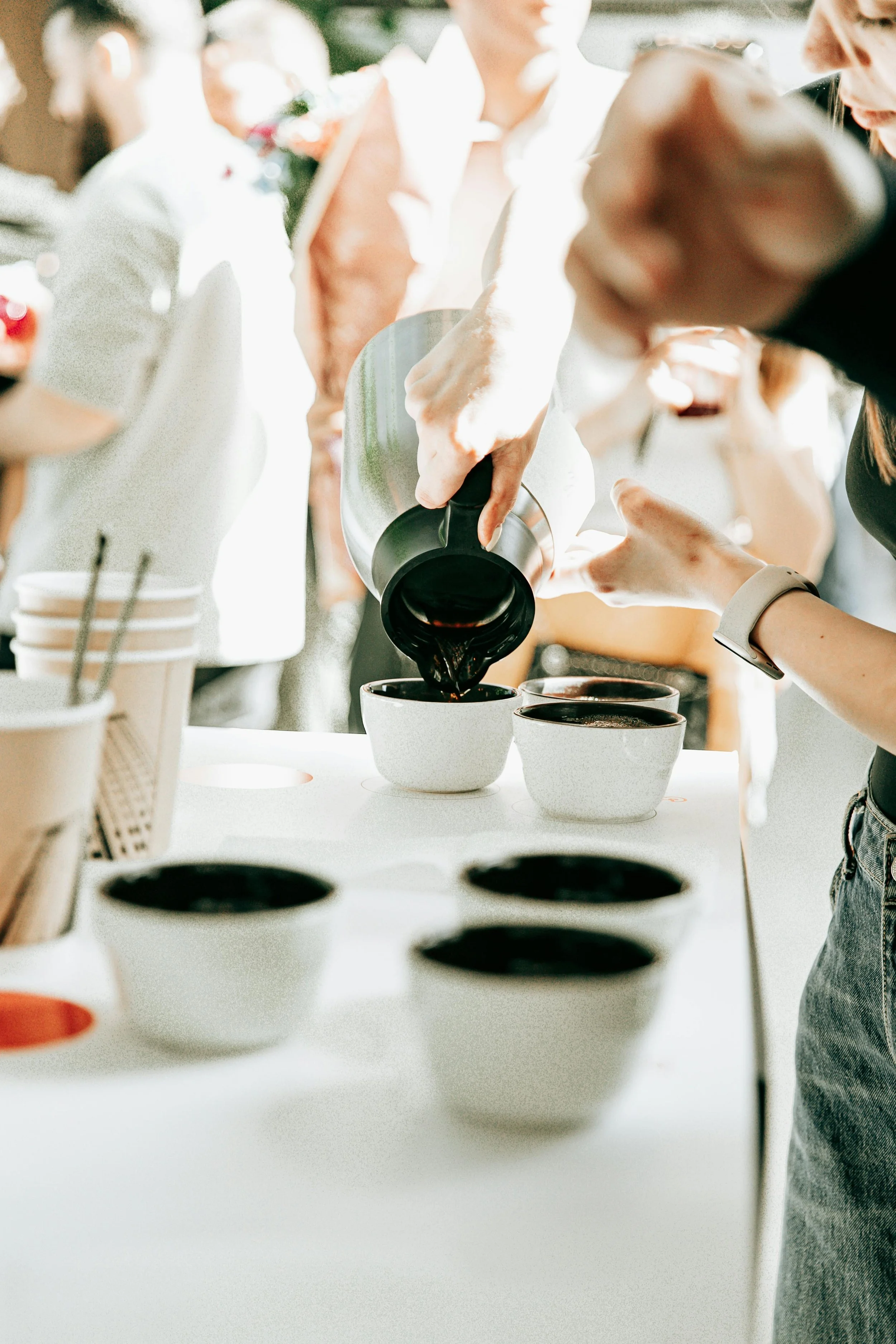 A person pouring coffee into cups at a gathering or event, with several cups on a white table and other people in the background.