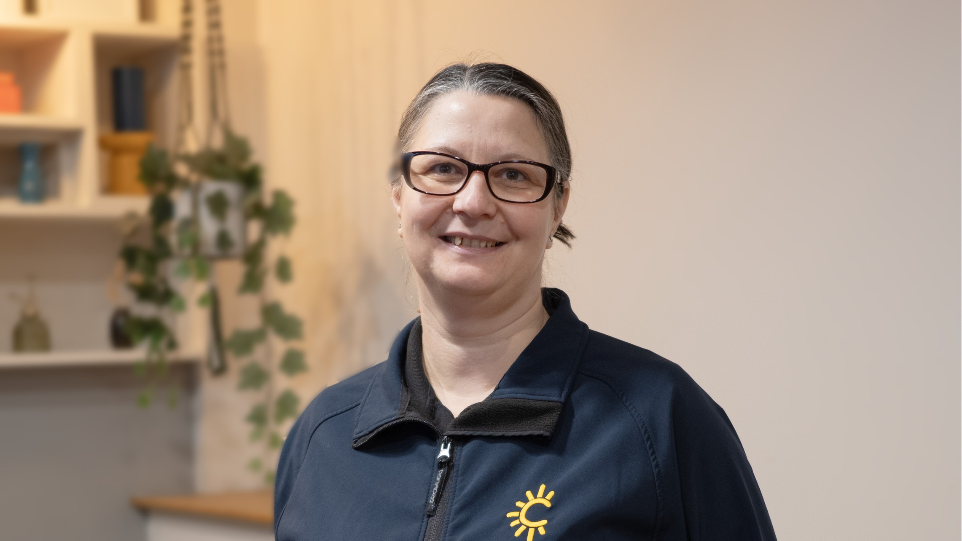 A member of Radiant Cleaners team with glasses smiling at the camera indoors, wearing a dark shirt with a yellow sun logo, in front of a blurred background with shelves and houseplants.