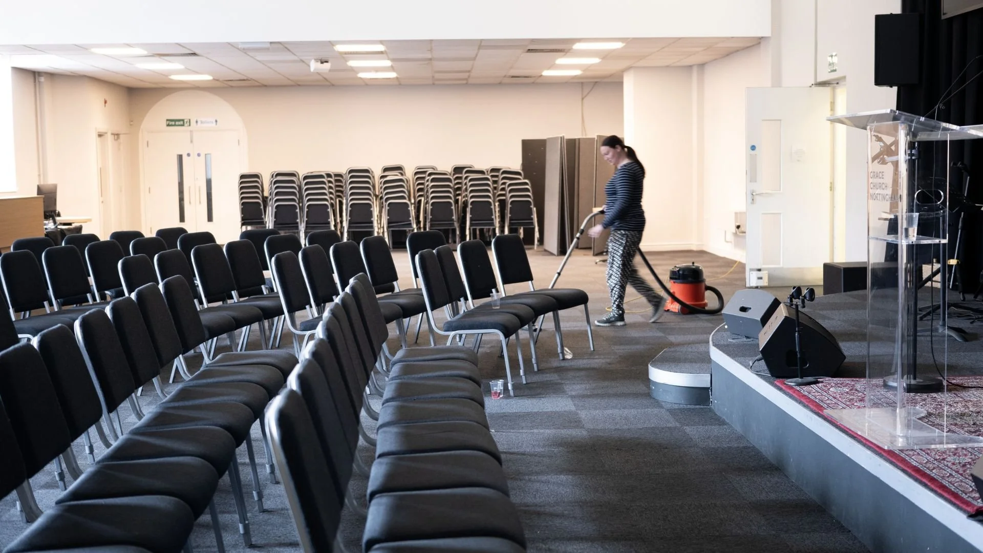 Empty chairs arranged in rows in a large room, with a woman vacuuming the floor near a stage and a clear podium on the right side.