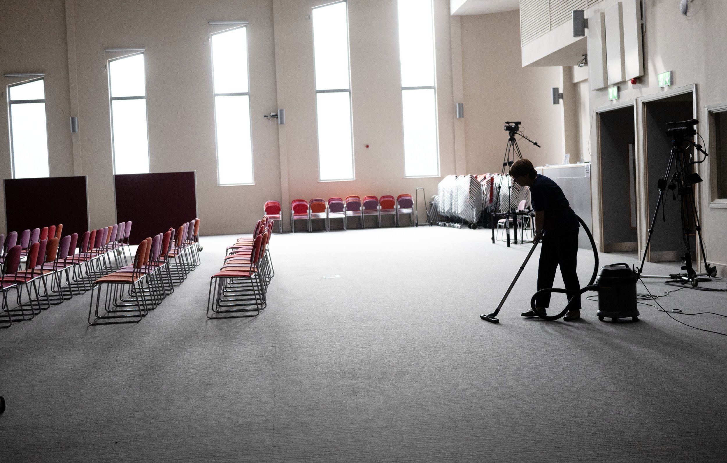 Empty chairs set up in a large, well-lit room with tall windows, with a person vacuuming the carpet near cameras and equipment, possibly for an event or filming.