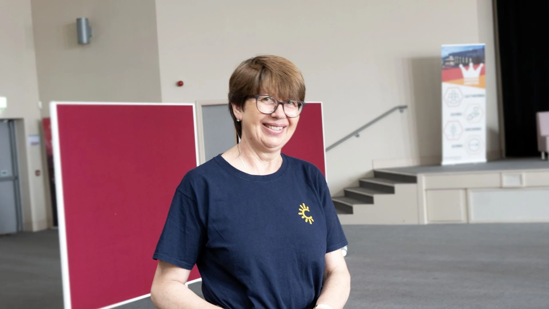 Smiling cleaner with short brown hair and glasses stands indoors in front of a red partition, wearing a navy Radiant Cleaners t-shirt.