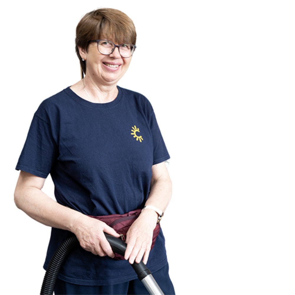 A smiling middle-aged woman with short brown hair, wearing glasses and a navy blue T-shirt with a sun logo, holding a vacuum cleaner.