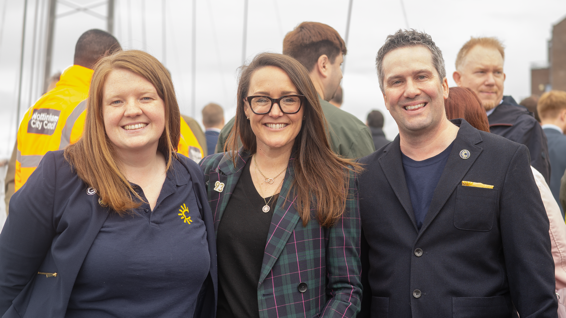 Three smiling individuals standing together at an outdoor event, with other people in the background, one wearing a yellow safety vest with 'Nottingham City Council' written on it.
