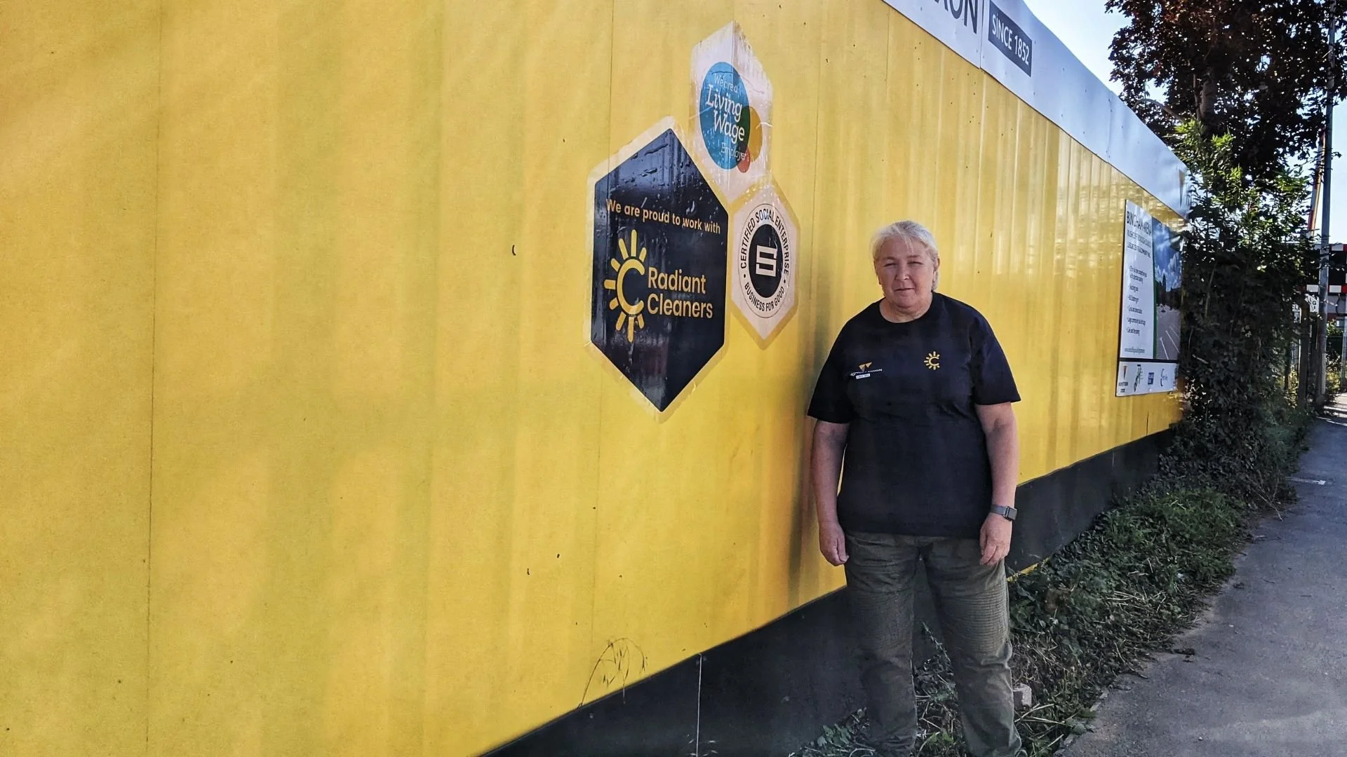 A woman with short gray hair standing next to a bright yellow wall with various logos and signs, including a 'Radiant Cleaners' logo and a 'We’re proud to work with' message.