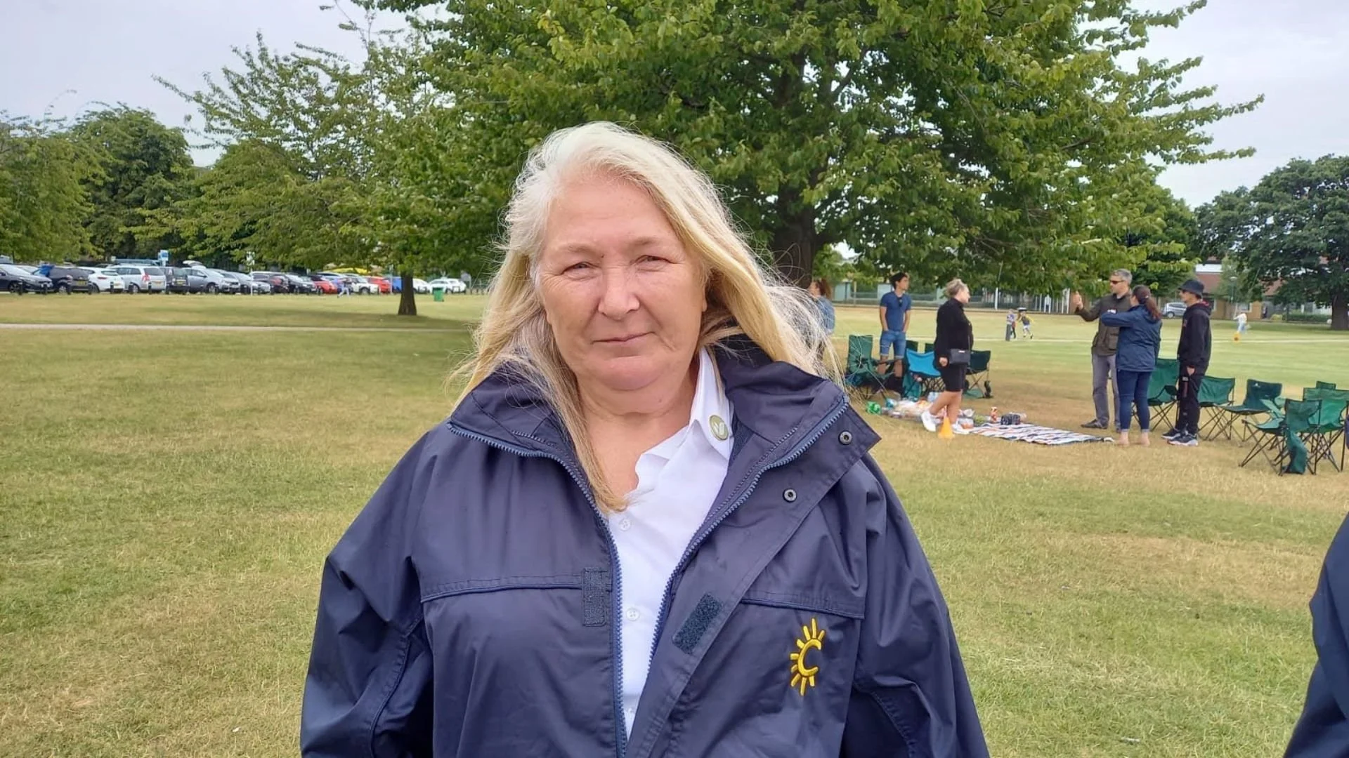 A woman with long blonde hair wearing a navy jacket and white shirt stands in a park on a cloudy day. In the background, several people are gathered near a green tree, some standing and talking, others sitting on folding chairs. There is a row of cars parked in the distance and a blanket with various items laid out on the grass.