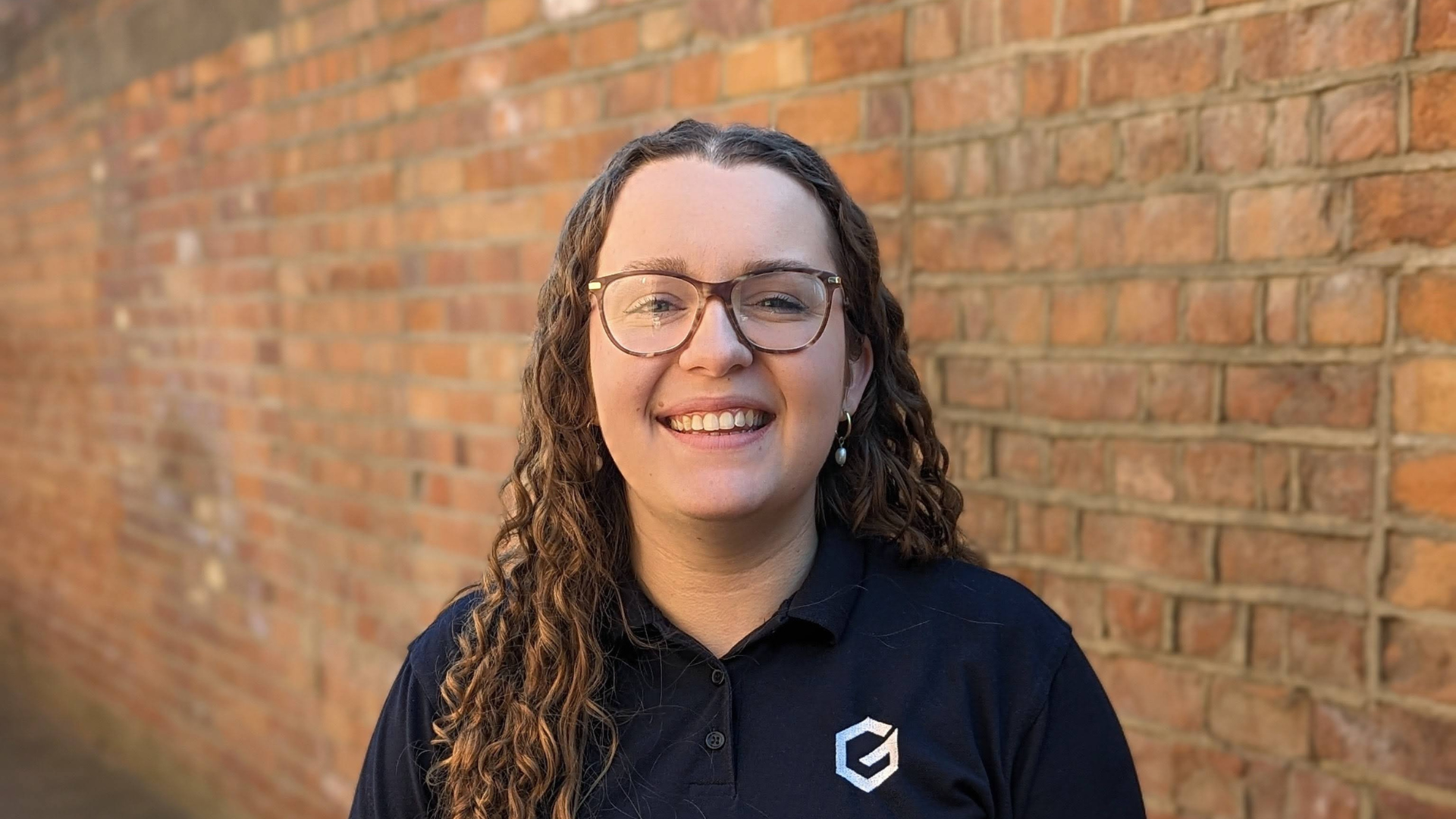 A young woman with curly hair, glasses, and earrings smiling in front of a brick wall.