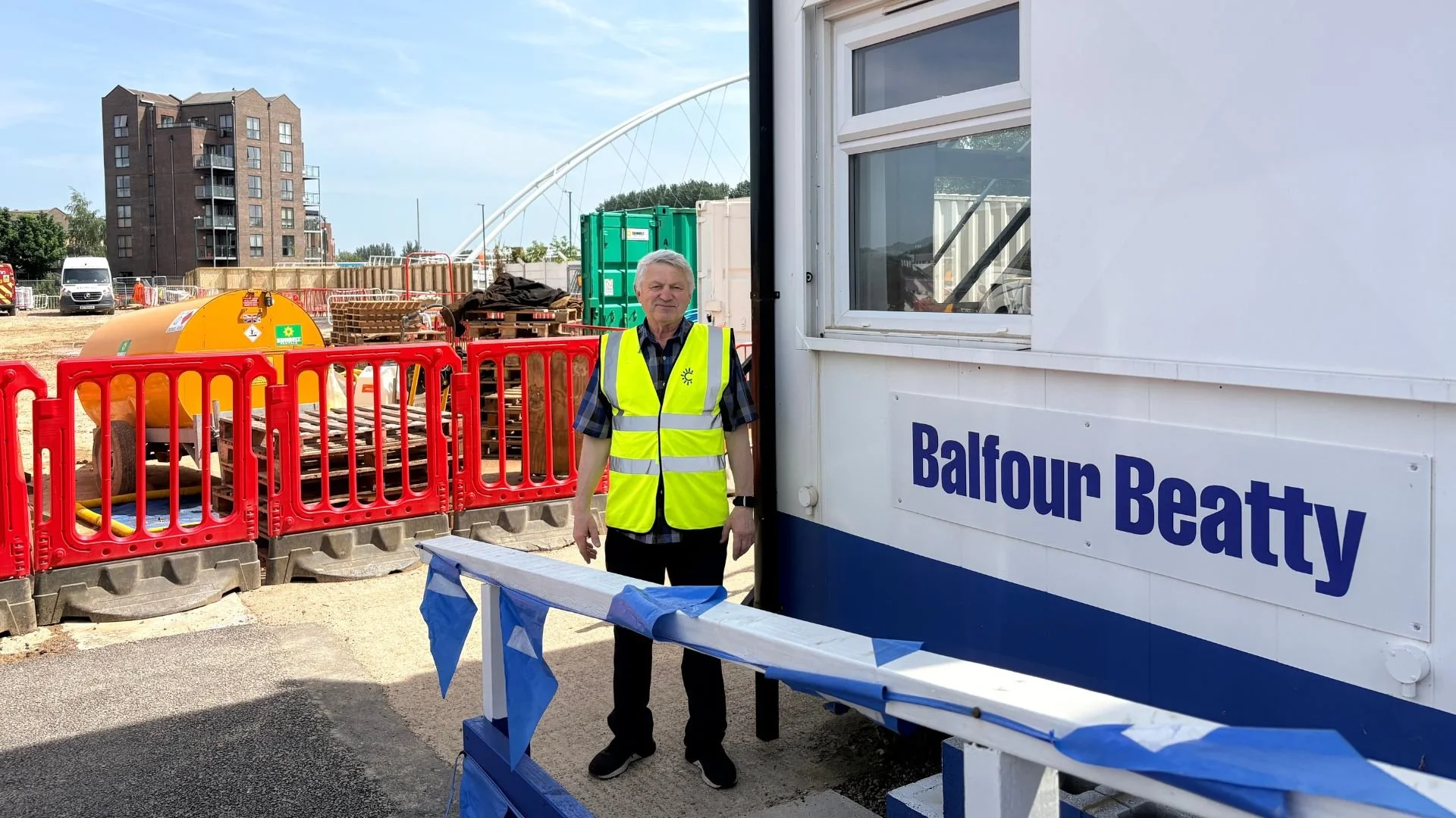 A man wearing a yellow safety vest standing next to a portable building with the label "Balfour Beatty" at a construction site. There are construction materials, temporary barriers, and a large orange water tank in the background.