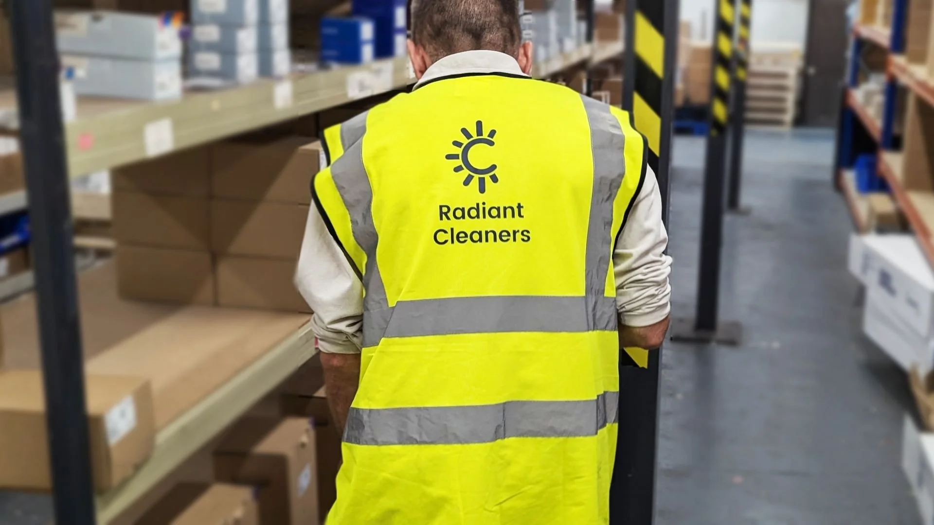 A worker in a warehouse wearing a bright yellow safety vest with the words "Radiant Cleaners" and a logo of a sun and a cleaning tool on the back, looking at shelves of boxes.