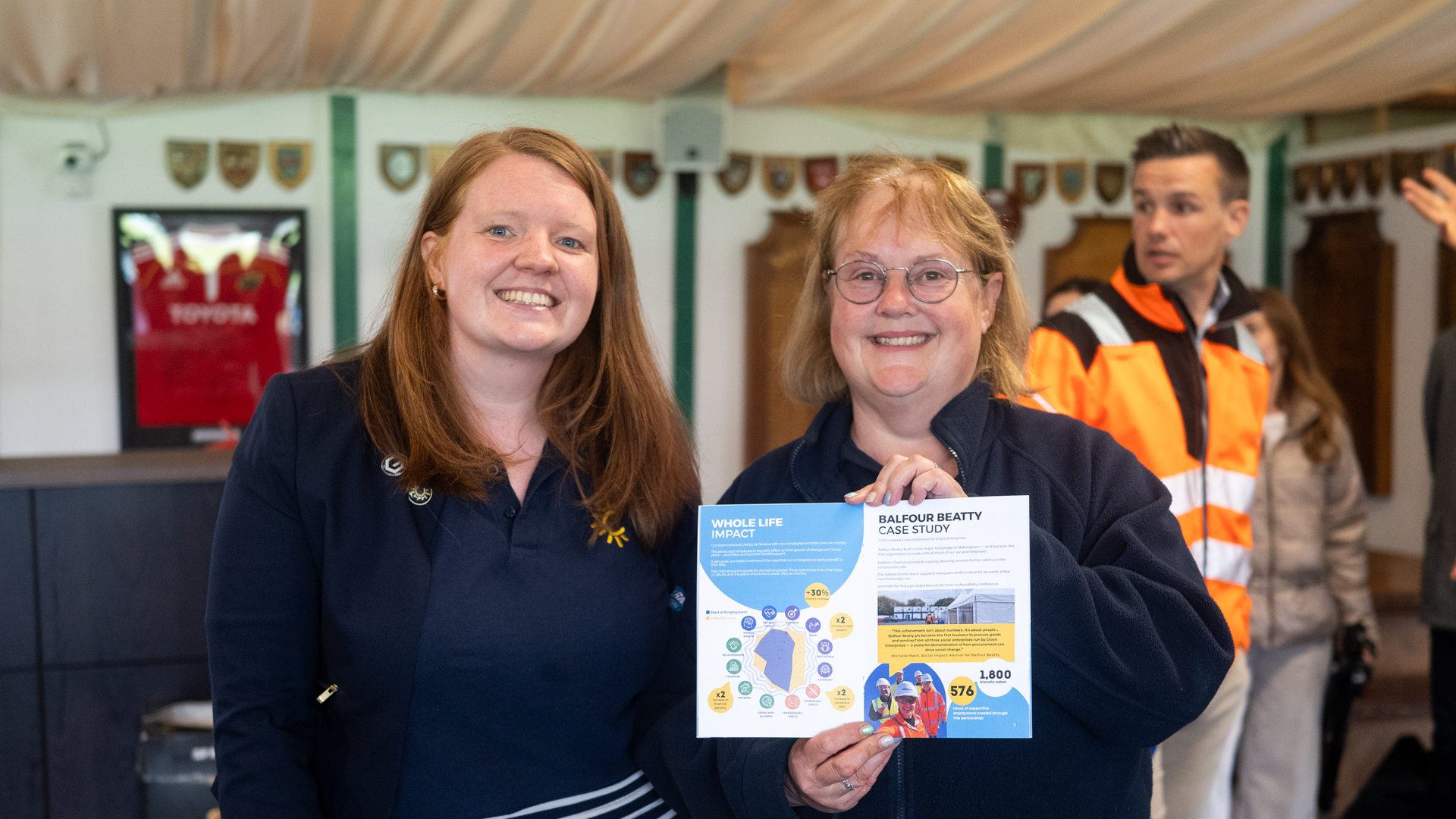 Two women smiling; one holding a fact sheet on Radiant Cleaners' social impact, with a man in a high-visibility jacket in the background.