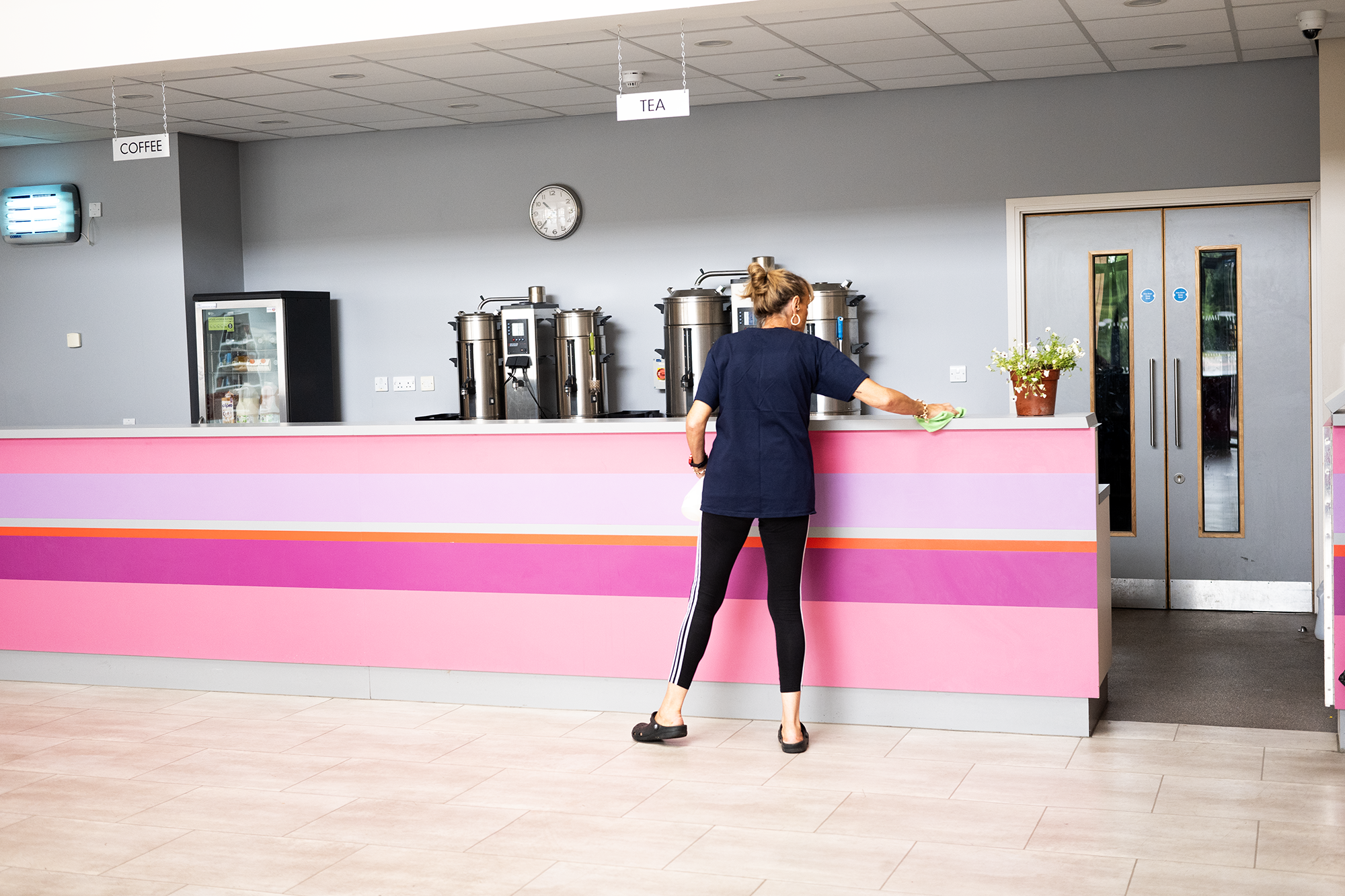 A woman cleaning the counter of a pink and purple coffee stand, with coffee and tea signs hanging above, a clock on the wall, and a glass door in the background.