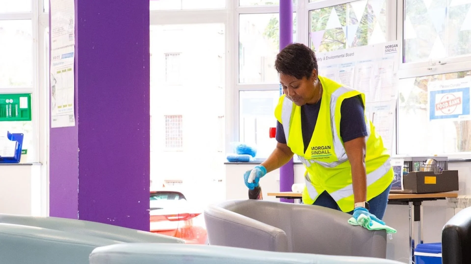 A woman wearing a yellow safety vest and gloves is cleaning a hospital bassinet in a bright room with purple and white walls, and large windows letting in natural light.