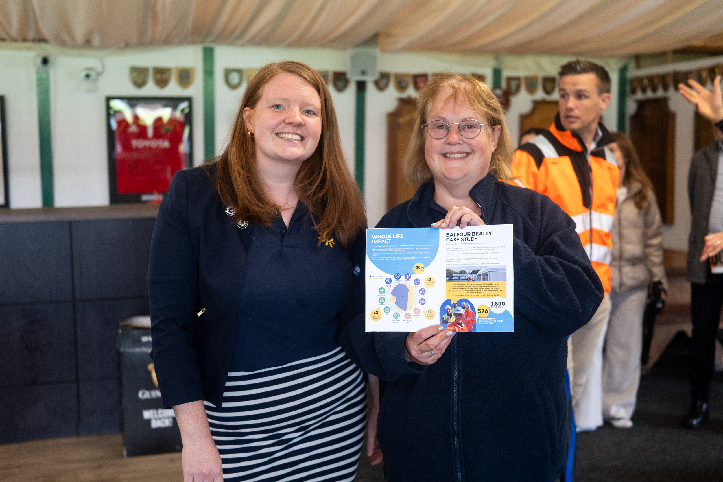 Two women smiling at the camera, one holding a flyer about Balfour Beatty case study, with a group of people in the background at an indoor event.