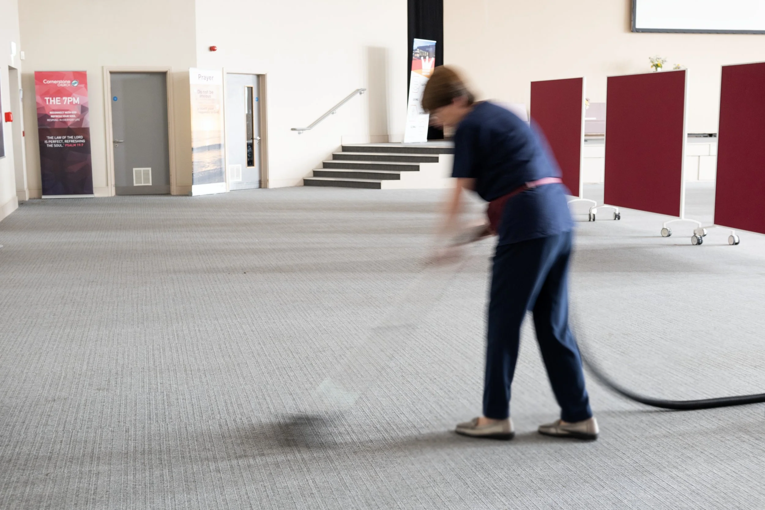 A person in blue clothing and white shoes is walking or sweeping on a beige carpet in a spacious indoor area. There are red and white privacy screens on wheels in the background, along with a set of stairs and a wall with posters and a door.