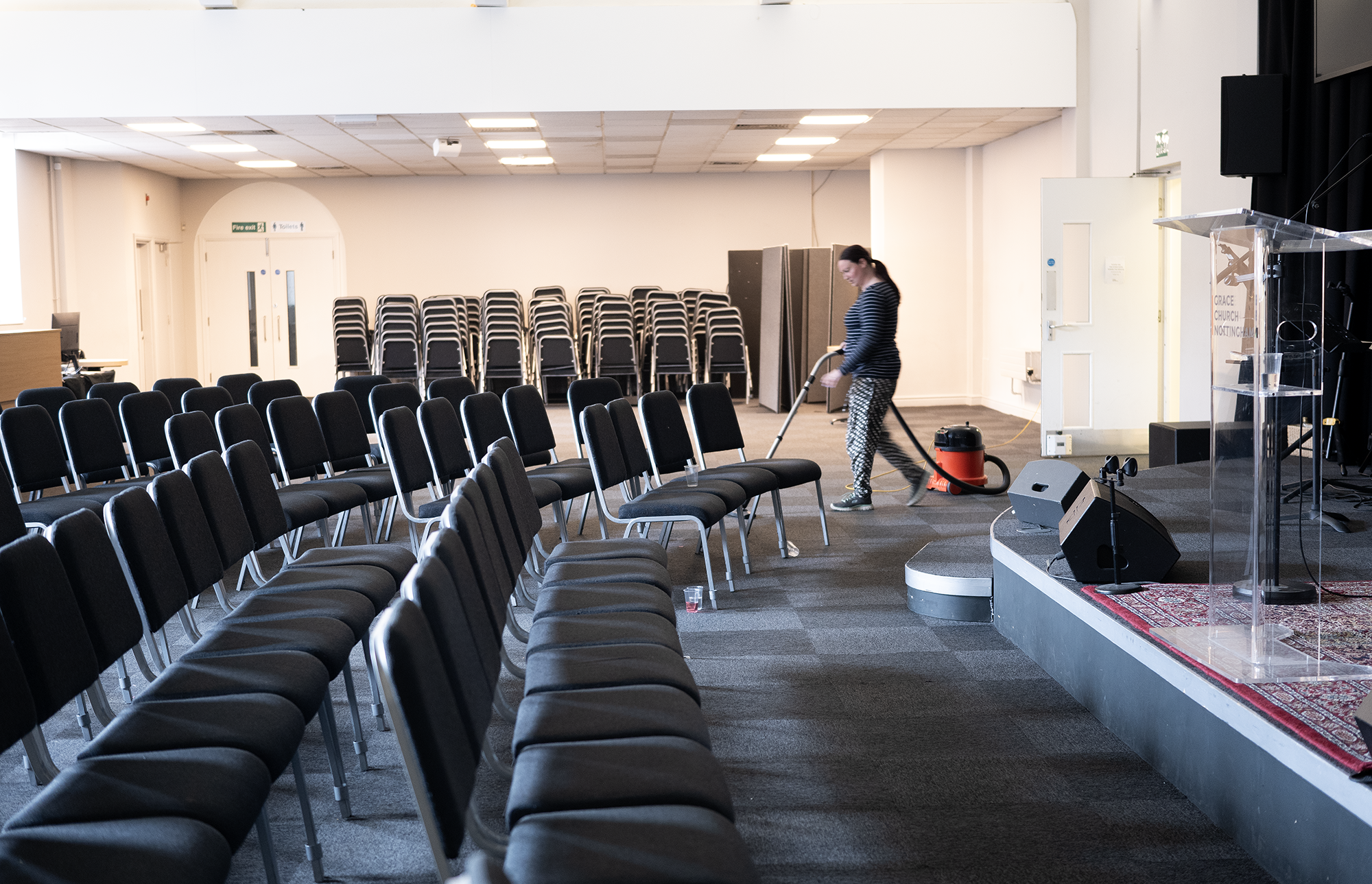 A woman cleaning the floor in an empty conference room with rows of black chairs, a stage with a clear podium, and stacked chairs in the background.